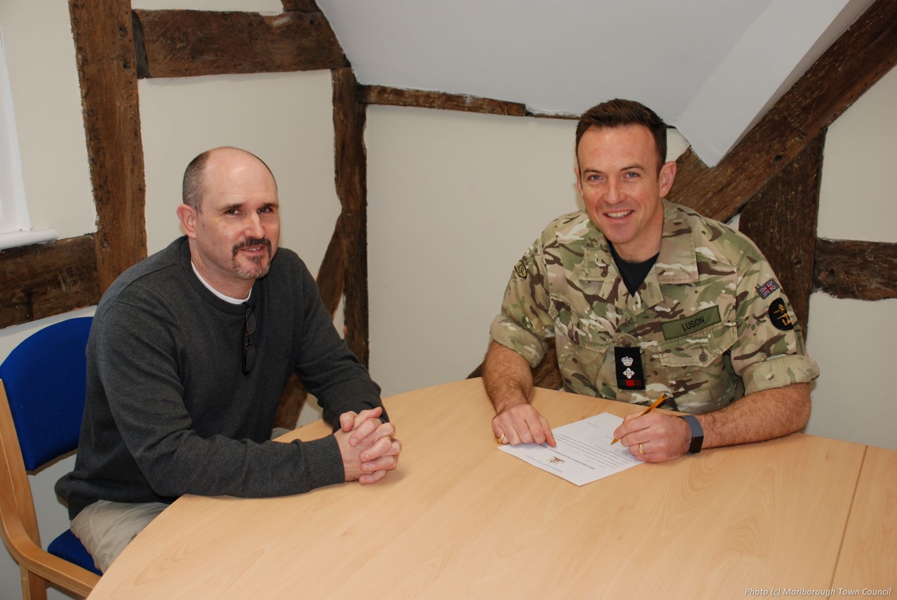 Two men sit at right angles at a table.  One is signing a document.  Both are smiling.