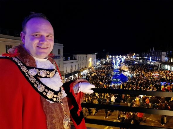A man wearing a red ceremonial robe and chain of office smiles at the camera.  He stands on a balcony and beyond is a street filled with people.  There are decorative lights above a market