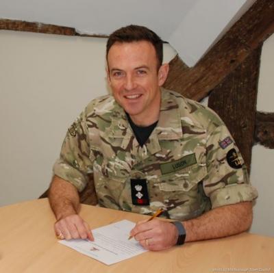 A man in uniform sits at a table with a piece of paper on it.  He is smiling at the camera while holding a pen in the act of signing the paper. 
