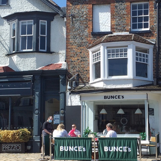 a photograph of people sitting at cafe tables on a pavement.  Surrounding them are canvas screens with the word Bunces written on it.  A brick and painted building is immediately behind with large win