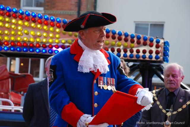 A man in a ceremonial blue coat and tricorn hat reads aloud from a scroll