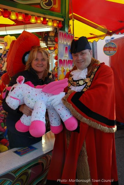 A man and woman smile at the camera.  They hold a large soft toy unicorn
