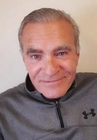 a head and shoulders photo of a mature man, smiling at the camera.  He is pale skinned, clean shaven with hair brushed back from his forehead.. He wears a grey zip up jumper.
