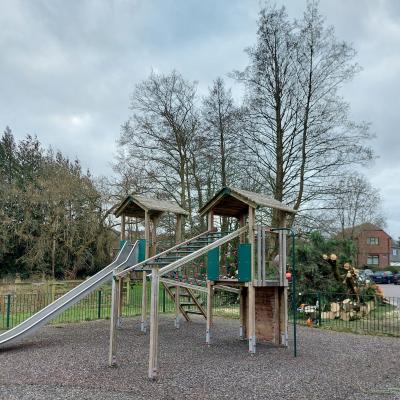 An outdoor scene of a children's play area. A wooden structure with slides attached stands in front of trees, some of which have been freshly felled