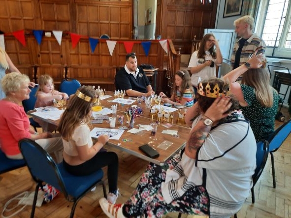 A photo of a room.  Adults and children sit around a table with craft materials, making crowns and colouring in pictures