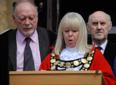a blonde haired woman looks down and is speaking, with sombre looking men standing behind
