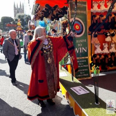 A woman in ceremonial dress holds a bow and arrow at a fairground stall