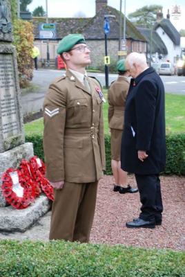a man in dark clothing stands with head bowed, before a poppy wreath at the foot of a memorial