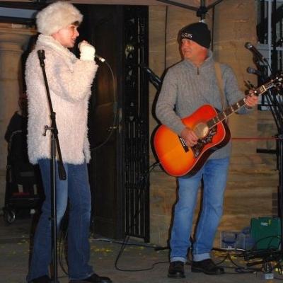 a woman wearing a white coat and hat sings into a microphone and a man plays guitar