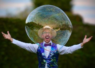 a man wearing a hat, waistcoat & bowtie and white shirt has his head in a large transparent bubble