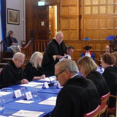 A man in a black robe stands, while other people sit around a table