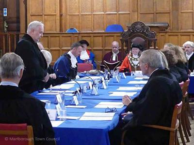 A man in black gown stands, speaking to people seated around a table
