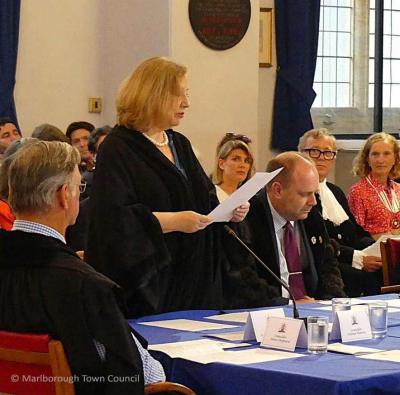 A woman with a fair bob wears a black gown.  She is reading from a sheet of paper