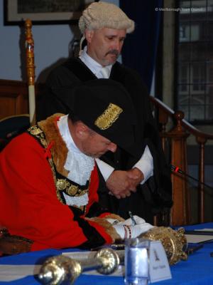 A man in a red robes of office is seated, signing a paper.  A man in a black gown wearing a horsehair wig stands, observing