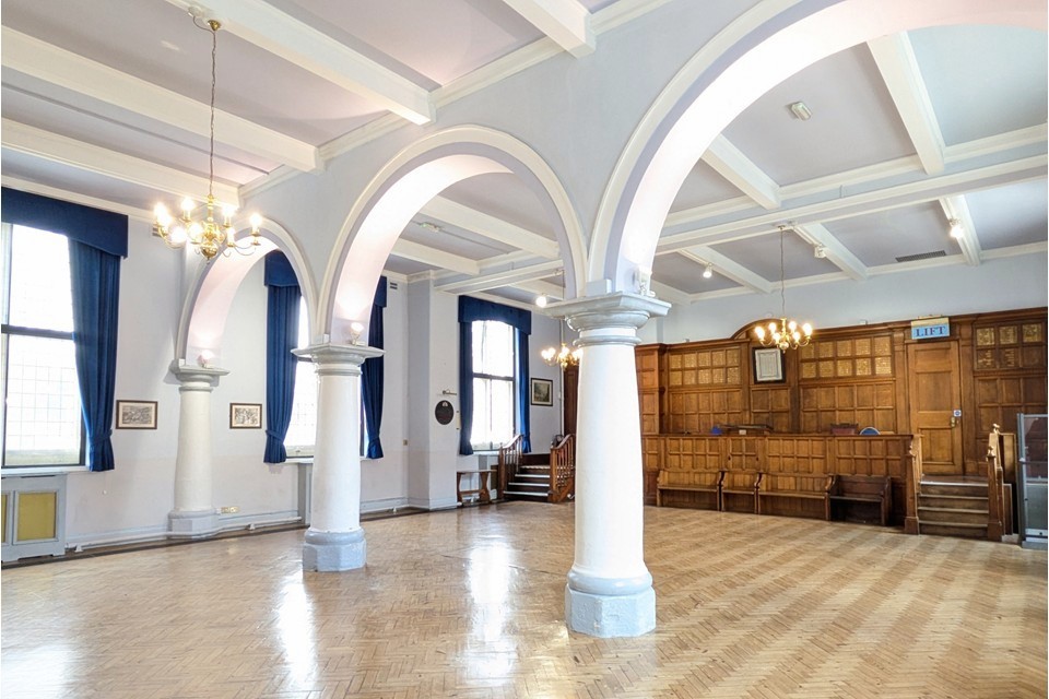 The Court Room A large bright empty room with large windows along either side. Pillars and arches bisect part and there is a wood panelled platform at one end